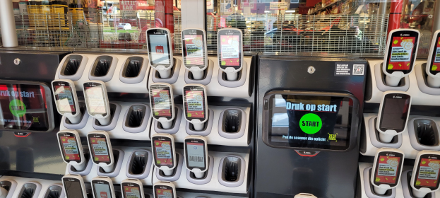 Ein Verkaufsautomat mit mehreren Handys auf der Oberseite, vor einem Hintergrund voller Menschen, Geschäfte, Schilder und Lichter.