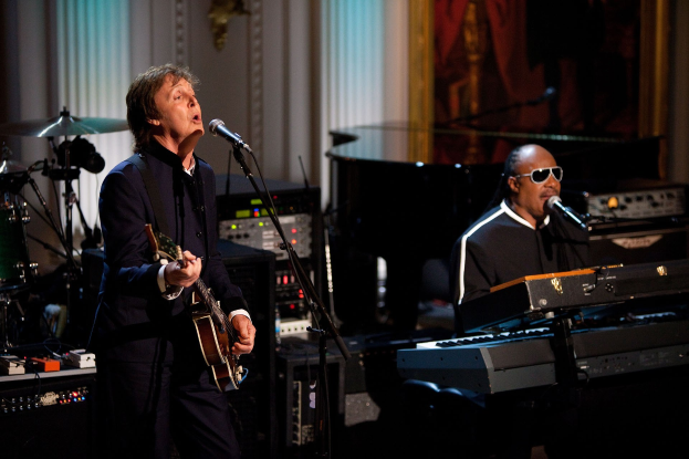Paul McCartney spielt Gitarre und singt in ein Mikrofon, während Stevie Wonder eine Sonnenbrille trägt und am Piano auf der Bühne der Grammys spielt, mit Instrumenten und einem gerahmten Bild im Hintergrund.
