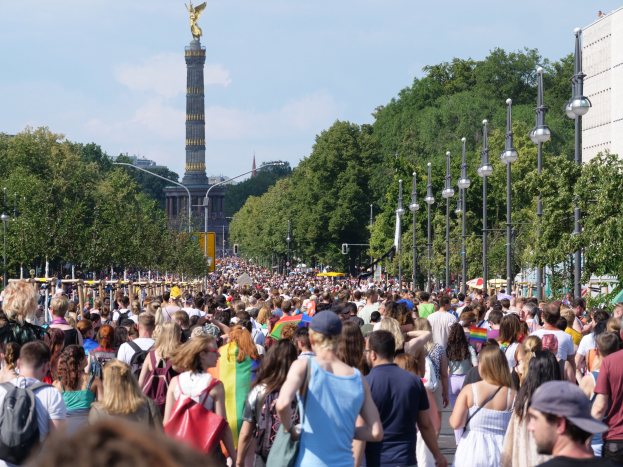 Eine große Menschenmenge geht eine Straße in Berlin, Deutschland, entlang, mit Laternenpfählen am Weg, Bäumen und einem Turm mit einer Statue im Hintergrund und Gebäuden unter einem bewölkten Himmel.