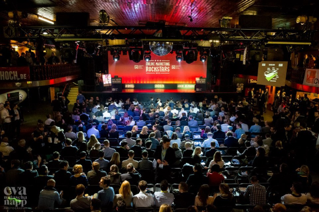 Eine große Zuschauermenge sitzt in Stühlen, die einer hell erleuchteten Bühne mit Bildschirmen und Bannern gegenüberstehen und auf ein Online-Marketing-Event warten.