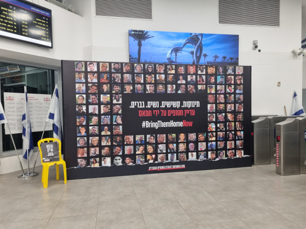 Eine große Wand zeigt zahlreiche Fotos, einen gelben Stuhl, israelische Regierungsflaggen auf Stangen, eine Tafel mit der Aufschrift "Bring Them Home Now", einen montierten Bildschirm, Deckenleuchten und andere Gegenstände.