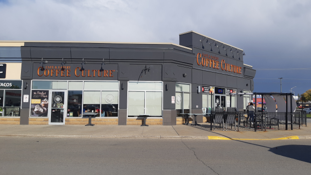 Ein Kaffeeladen an einer Straßenecke mit einer Straße im Vordergrund, einem Gebäude im Hintergrund, einigen Fahrzeugen, Bäumen und einem blauen und weißen Himmel.