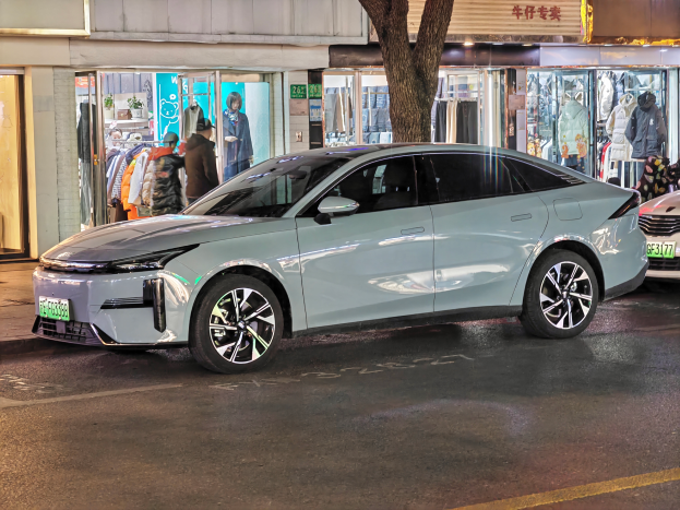 Ein Mitsubishi Lancer EV-Elektroauto, das nachts auf einer Straße geparkt ist, umgeben von Gebäuden, Geschäften, Schaufensterpuppen mit Kleidern, Schildern mit Text, Lichtern und anderen Objekten.