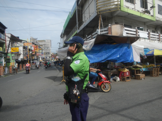 Ein Polizeibeamter in grüner und blauer Uniform mit einer blauen Mütze steht auf einer Straße und spricht auf einem Handy, während Fahrzeuge vorbeifahren und Marktstände rechts von ihm sind, unter einem bewölkten Himmel.