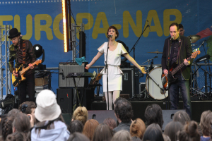 Eine Bühne mit vier Musikern: Eine Frau in einem weißen Kleid singt in der Mitte, zwei Männer spielen Gitarren auf beiden Seiten und eine Menschenmenge und Lautsprecher im Hintergrund.