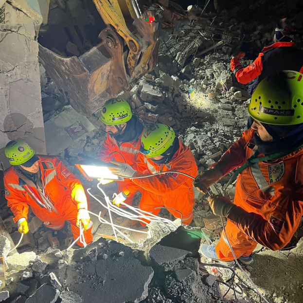 Gruppe von Menschen in orangefarbenen Uniformen und grünen Helmen bei der Arbeit an Schutt mit einem Kran im Hintergrund während einer Rettungsaktion nach einem Einsturz.