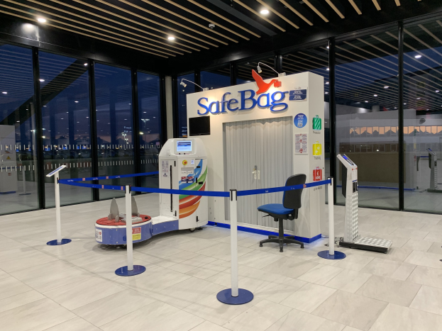 Sicherheitskontrolle am Flughafen mit Stuhl, Absperrpfosten, Maschinen, Bildschirm, bedruckter Tafel, Glaswänden, Deckenleuchten und sichtbarem Boden.