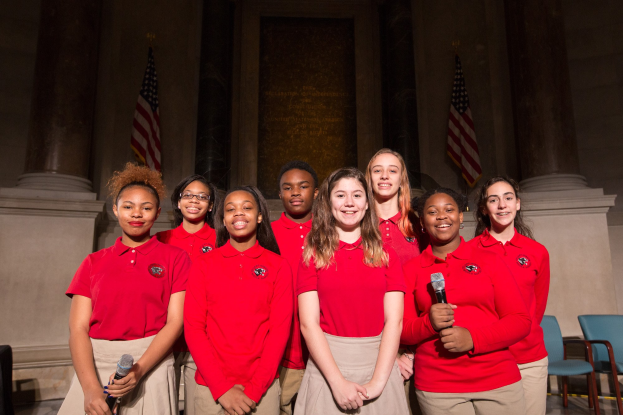 Gruppe junger Frauen in roten Hemden, die strahlend Mikrofone halten und vor Stühlen, Säulen und Fahnen stehen, wahrscheinlich Teil eines nationalen Jugendchors.