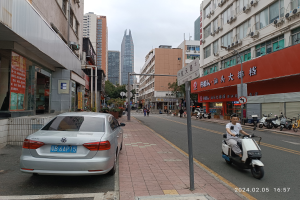 Ein Mann fährt mit einem Roller eine Straße entlang, die von hohen Gebäuden gesäumt ist. Menschen gehen auf den Gehwegen, Fahrzeuge fahren auf der Straße und Bäume sind unter einem klaren blauen Himmel zu sehen.