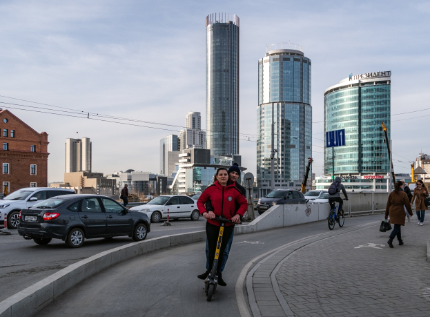 Ein Mann in einer roten Jacke und schwarzen Hosen fährt auf einem Elektroroller die Straße entlang, umgeben von Fußgängern und Radfahrern unter einem bewölkten Himmel.