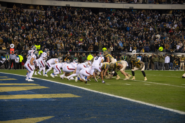 Ein Fußballspiel auf einem Stadionfeld mit Spielern in Sportkleidung und Helmen und Zuschauern im Hintergrund.