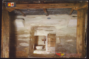 Ein altes Foto einer Grabkammer mit einer hölzernen Tür, einem Kreuz an der Wand, einem Dach oben und Text mit einem Logo unten.