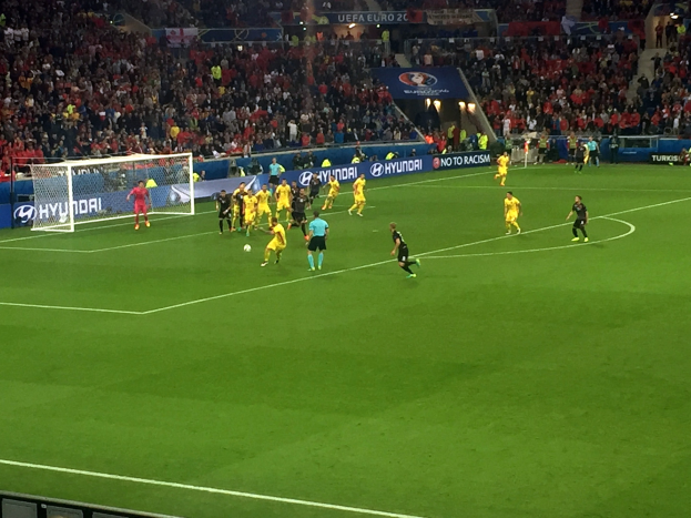 Fußballspieler, die auf einem Feld in einem Stadion mit einem Netz auf der linken Seite spielen, Zuschauern in den Rängen und beleuchteten Tafeln im Hintergrund.