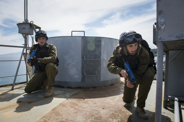 Zwei israelische Soldaten in Uniform, Helmen und Schutzbrillen stehen auf dem Deck eines Schiffs und halten Gewehre, mit einem Panzer, Metallstangen, Wasser, Hügeln und einem bewölkten Himmel im Hintergrund.