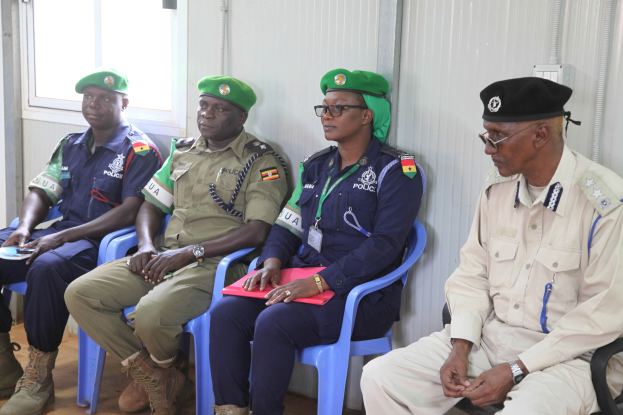 Vier Polizisten in Uniform und Mützen sitzen auf Stühlen während einer Schulung in einem Raum mit einer Wand und einem Fenster im Hintergrund, einer hält ein Buch und einen Stift.