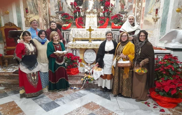 Eine Gruppe von Menschen steht vor einer Kirche während einer Weihnachtsfeier, mit Körben und Tellern in den Händen, Blumensträußen, Statuen, Kerzen und Gemälden im Hintergrund.