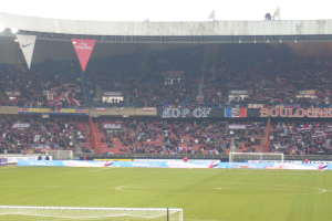 Ein Rasenplatz mit Torpfosten auf beiden Seiten, gefüllt mit Zuschauern im Hintergrund und zahlreichen Werbetafeln in der Mitte.