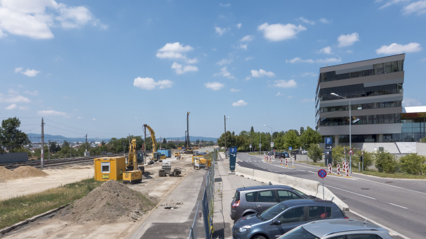 Eine Baustelle mit parkenden Autos, Gebäuden, Straßenlaternen, Strommasten, Schildern, Absperrpoller, Bäumen, Pflanzen, Gras, Sand, Kränen, Bahnschienen, Strommasten mit Drähten, Hügeln und einem bewölkten Himmel im Hintergrund.