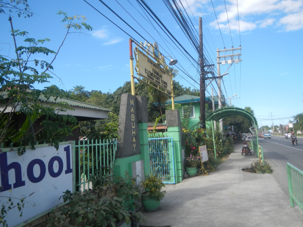 Schuleingang mit einem "Schule"-Schild am Straßenrand, Fahrzeugen, einem Bogen, einem Tor, einer Namensplakette, Pflanzen, Bäumen, Strommasten mit Drähten und einem bewölkten Himmel im Hintergrund.
