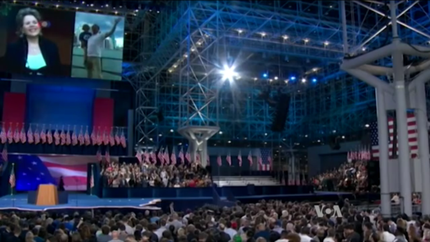 Eine große Menschenmenge sitzt vor einer Bühne mit einem Rednerpult, Mikrofon, Flaggen, einem Bildschirm und Beleuchtungsausrüstung und wartet auf eine Präsidentenrede.