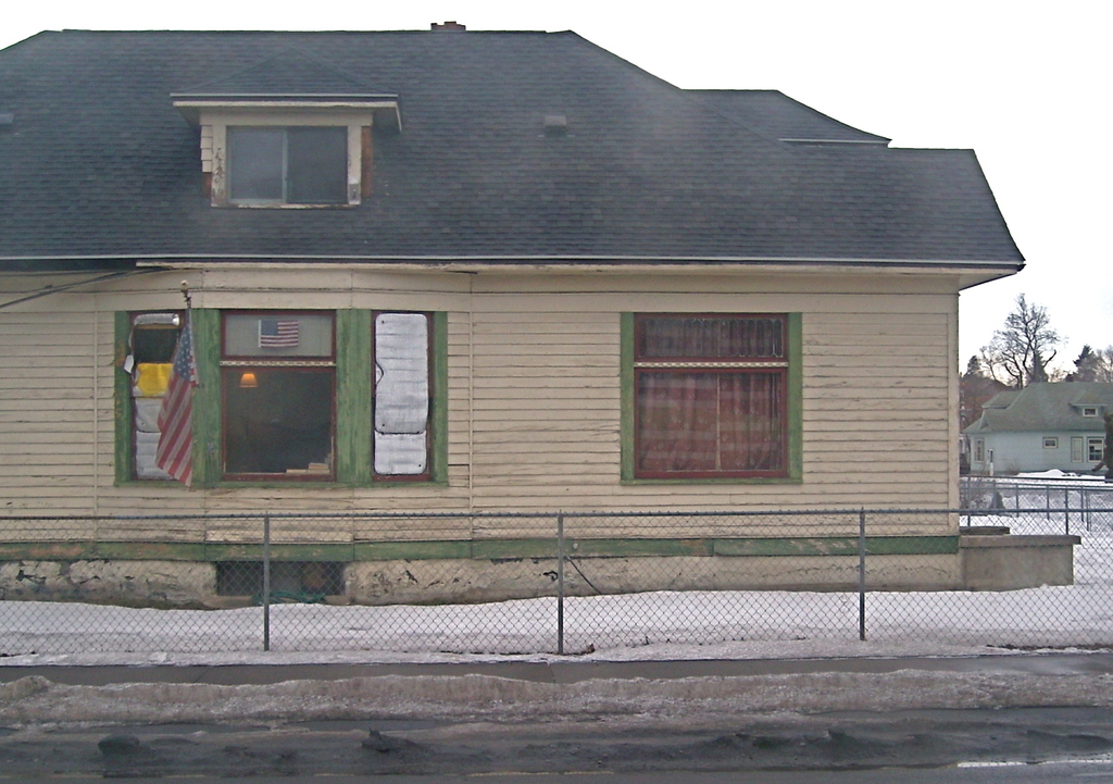 Ein Zaun ist in der Nähe einer schneebedeckten Fläche auf dem Boden positioniert, im Hintergrund ein Haus mit Dach und Glasfenstern sowie anderen Häusern, Bäumen und dem Himmel.