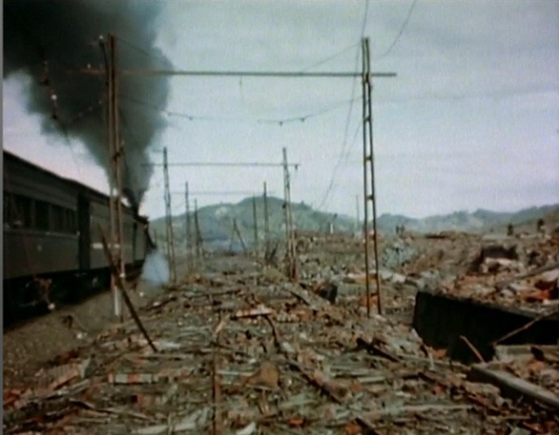Ein Zug fährt durch ein beschädigtes Gebiet mit Rauch, der aus ihm aufsteigt, umgeben von Strommasten und Drähten, mit Hügeln und einem klaren Himmel im Hintergrund und verstreuten Trümmern auf dem Boden.