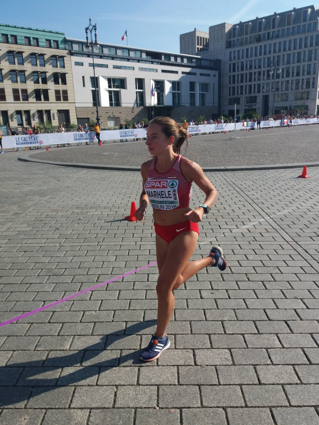 Eine Frau in einem Sportdress läuft durch eine Marathon-Strecke, die von Hütchen, einer Schleife und einem Banner markiert ist, mit Zuschauern und Fahnen entlang der Straße unter einem bewölkten Himmel.