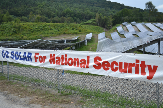 Ein Zaun mit einer Aufschrift "Gehen Sie Solar für die nationale Sicherheit" vor einem Solarpanel-Feld, Bäumen und Gras unter einem klaren blauen Himmel.