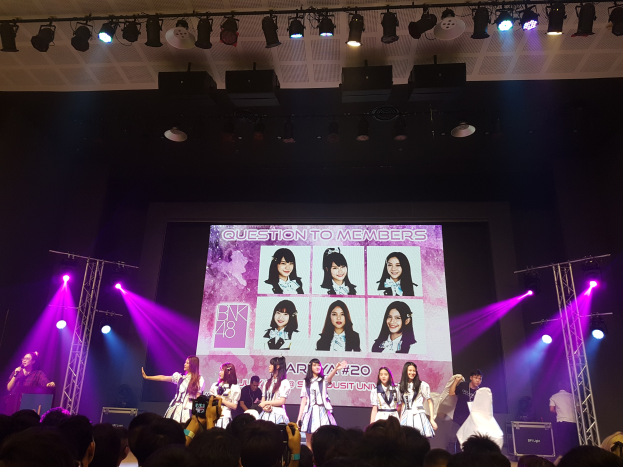 Eine Bühnenperformance mit einer K-Pop-Gruppe BK48, bei der Mitglieder auf der Bühne stehen, Mikrofone halten und Instrumente spielen, während unten eine Menge Fotos macht; helle Lichter und Träger umgeben die Szene, und ein großer Bildschirm zeigt BK48-Mitglieder im Hintergrund.