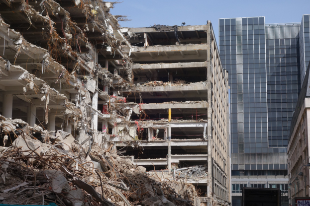 Ein großes Gebäude in einer städtischen Umgebung abgerissen, umgeben von intakten Strukturen, Straßeninfrastruktur, Fahrzeugen, Bäumen und einem klaren Himmel.