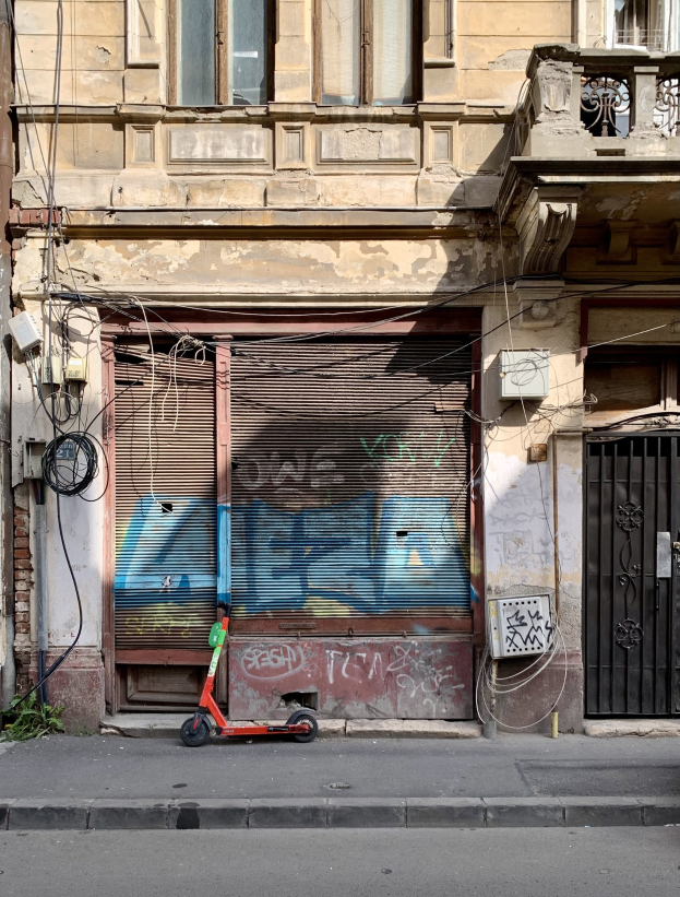 Ein roter Roller, der auf der Straße neben einem Gebäude mit Fenstern, Rollläden und einem Tor geparkt ist, mit sichtbaren Kabeln und anderen Gegenständen entlang der Straße.