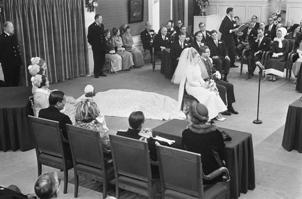 Schwarz-weiß-Foto einer Hochzeitszeremonie mit sitzenden und stehenden Menschen, einem Mann mit Mikrofon, einer Frau mit Blumenstrauß, einem Podium mit Mikrofon auf der rechten Seite und Blumenschmuck und Vorhang auf der linken Seite.