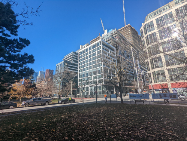 Eine Stadtstraße in Toronto mit hohen Gebäuden, fahrenden Fahrzeugen, Fußgängern auf dem Gehweg, Bäumen und verstreuten trockenen Blättern auf dem Boden unter einem sichtbaren Himmel.