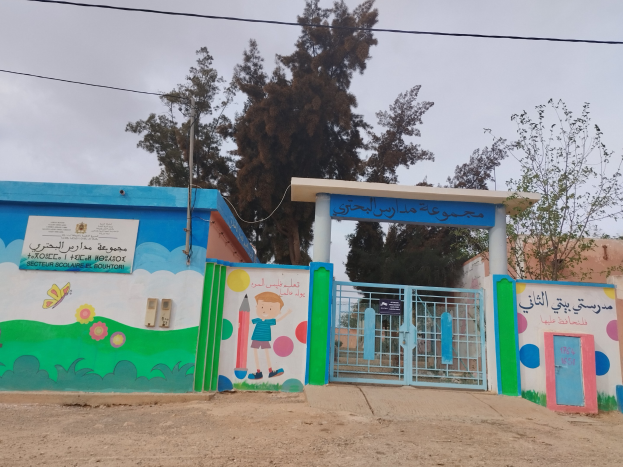 Blau-weißes Gebäude mit einer Wandmalerei, einem Tor und einer Namensplakette, das als Eingang zu einer Schule für Kinder dient, umgeben von Häusern, Bäumen, einem Strommast mit Drähten und einem bewölkten Himmel.