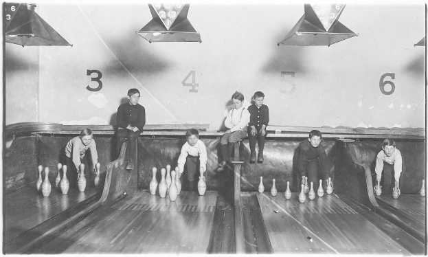 Schwarzes Foto von Kindern, die Bowling in einer Bahn mit aufgestellten Kegeln und nummerierter Wand im Hintergrund spielen.