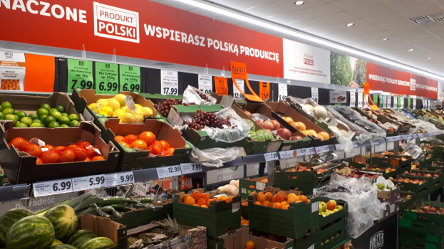 Ladeninneres mit verschiedenen Obst- und Gemüsesorten in Kartons mit Preisschildern, Wandtafeln mit Text und Bildern sowie Deckenbeleuchtung.