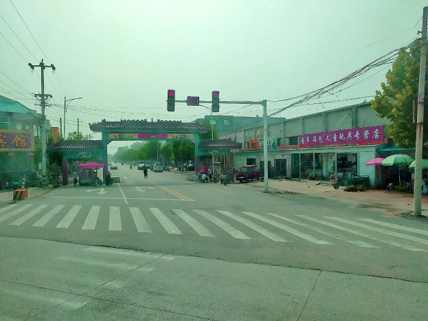 Stadtstraßenkreuzung mit Zebrastreifen, Ampel, Fahrzeugen, Strommasten, Gebäuden, Beschilderung, Bäumen, Schirmen und ein Bogen im Hintergrund unter einem sichtbaren Himmel.