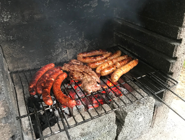 Grill mit Würstchen und Fleisch darauf, umgeben von glühender Kohle und Flammen, im Freien auf Gras stehend.