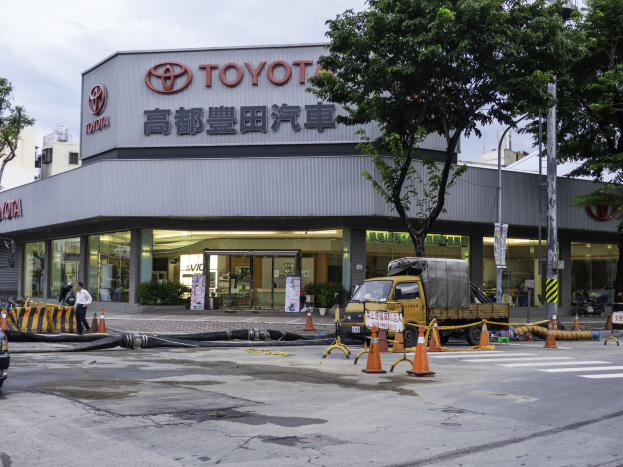 Ein Toyota-Autohaus in Tokio, Japan, mit Fahrzeugen, Verkehrskegeln, Rohren, Menschen, Bäumen, Pfählen, Brettern und Gebäuden im Vordergrund und einem bewölkten Himmel im Hintergrund.