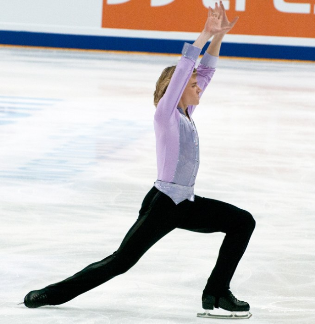 Ein Mann in einem lila Hemd und schwarzen Hosen führt eine elegantes Eiskunstlauf-Programm auf einem Eisstadion aus.