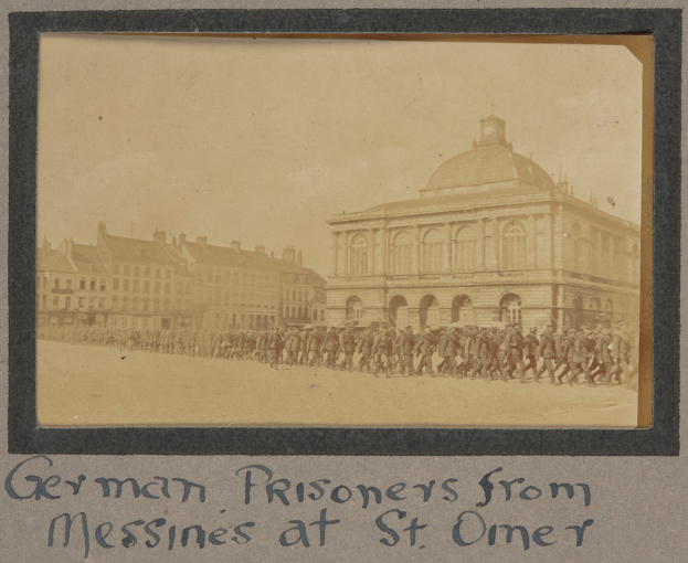 Deutsche Kriegsgefangene aus Messines vor einem Gebäude unter einem klaren blauen Himmel, mit Text am unteren Bildrand.