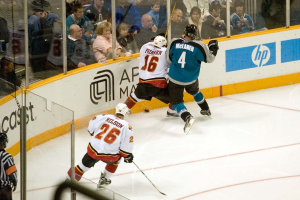 Ein Mann in einem sechzehn Nummern Jersey, Helm, Shorts und Schuhen steht neben einem anderen Mann in einer blauen Jacke, Brille, Helm und Hose neben einem Fenster, mit einem Publikum, das oben sitzt und einem Mann in einem vierundzwanzig Nummern Jersey, der einen Stock hält, unten, einem Schiedsrichter hinter Glas links unten und einem Werbeplakat rechts.
