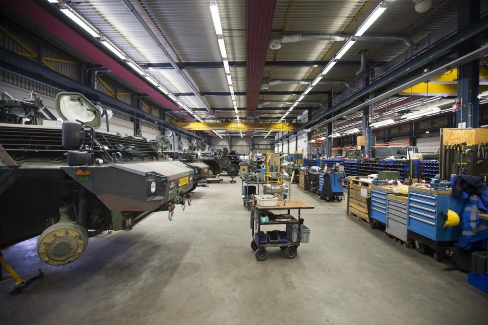 Großes Lager mit Maschinen, Fahrzeugen auf der linken Seite, Transportwagen mit Gegenständen und Holzboxen auf der rechten Seite sowie Deckenbeleuchtung.