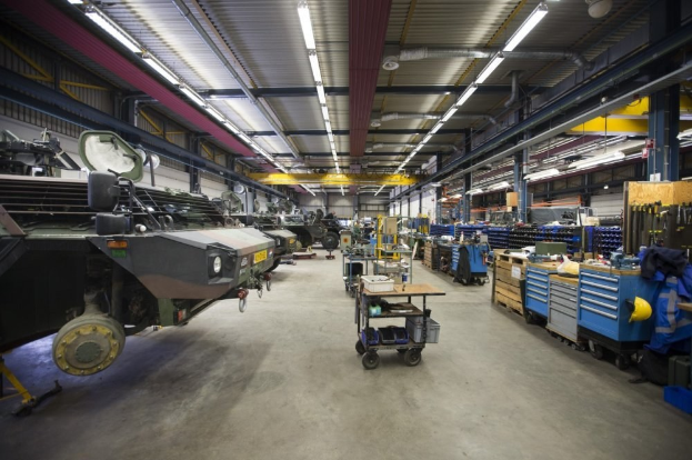 Großes Lager mit Maschinen, Fahrzeugen auf der linken Seite, Transportwagen mit Gegenständen und Holzboxen auf der rechten Seite sowie Deckenbeleuchtung.