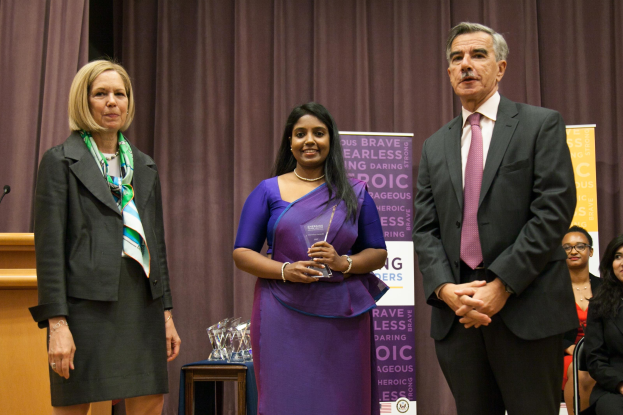 Zwei Frauen und ein Mann auf einer Bühne, wobei die Frau in der Mitte einen Award in einem lila Kleid hält, der Mann in einem schwarzen Blazer und pinker Krawatte, ein brauner Vorhanghintergrund, ein Podium mit Mikrofon links und Awards auf einem Tisch davor.