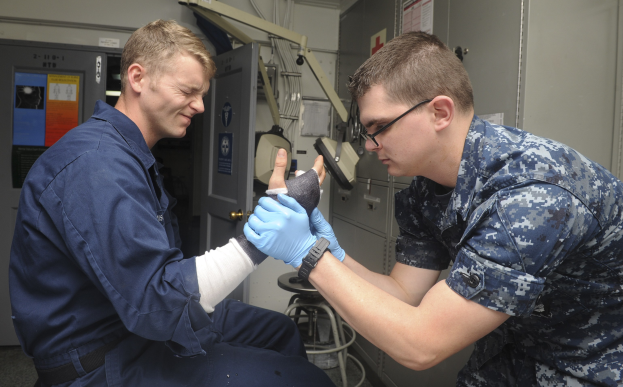 Zwei Männer in militärischer Uniform, einer sitzt in einem Stuhl mit einem Gips am Arm und der andere steht neben ihm, in einer medizinischen Einrichtung mit medizinischer Ausrüstung im Hintergrund.