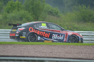 Ein Auto auf einer Rennstrecke mit Gras, einem Geländer und Bäumen im Hintergrund, mit Text und Zahlen darauf.