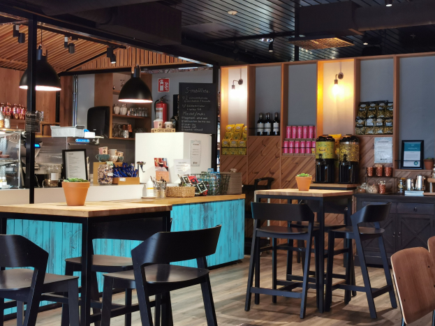 Inneneinrichtung eines Restaurants mit Holztischen, Stühlen, einer Topfpflanze auf einem Tisch, einer Schiefertafelmenü, einem Kühlschrank, einem Feuerlöscher, Regalen mit Flaschen, Schränken mit Gegenständen, einem gerahmten Foto an der Wand und Deckenleuchten.