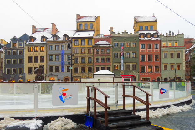 Eislaufbahn in einem Stadtplatz umgeben von Geb├Ąuden, Treppen, Gel├Ąndern, Informationsschildern, schneebedecktem Boden, Laternen, B├Ąumen und Himmel.