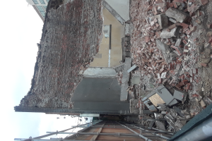 Ein teilweise abgerissener Bau mit verstreuten Ziegeln und Metallstangen, mit anderen Gebäuden und einem klaren blauen Himmel im Hintergrund.